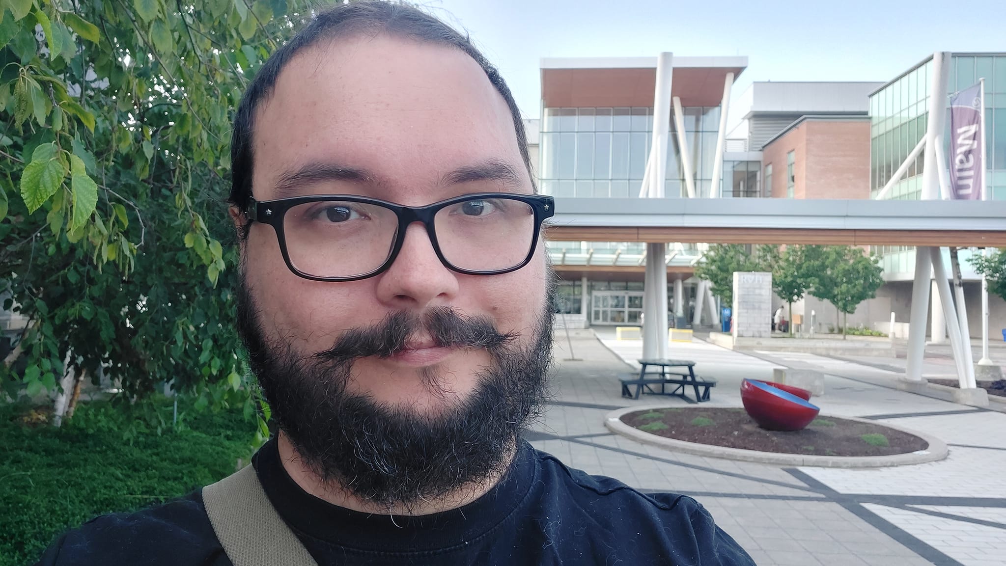 A man short hair and bushy facial hair stands in front of a brick and glass hospital building